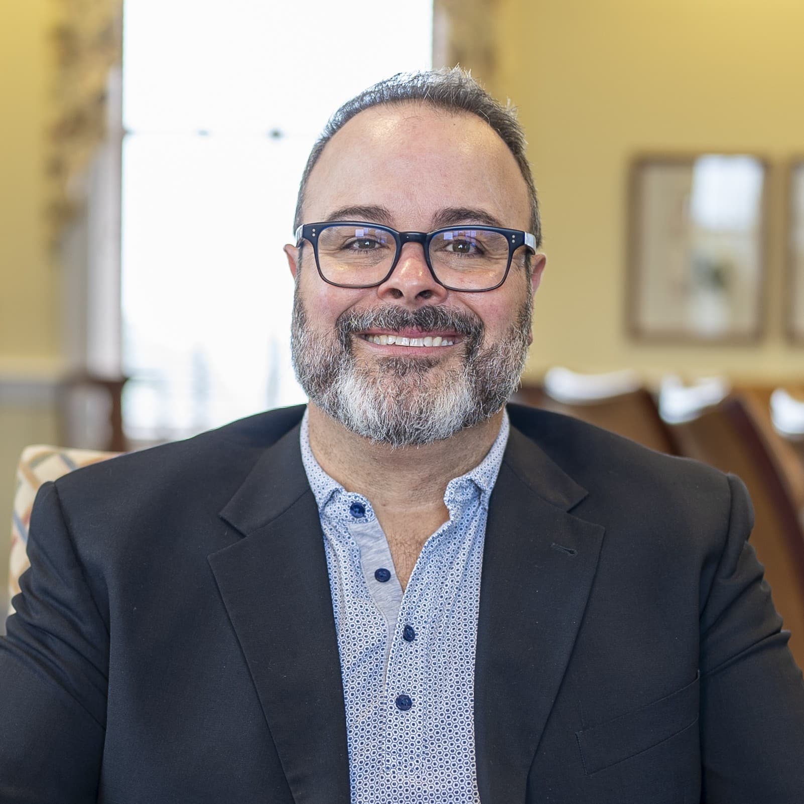 Smiling man with glasses, short beard, and mustache wears dark blazer over patterned shirt, sits indoors with blurred background.