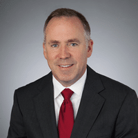 A man wearing a dark suit, white shirt, and red tie smiles confidently against a simple gray background.