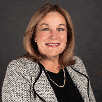 A woman with shoulder-length light brown hair in a grey textured blazer, black top, and pearl necklace smiles at the camera.
