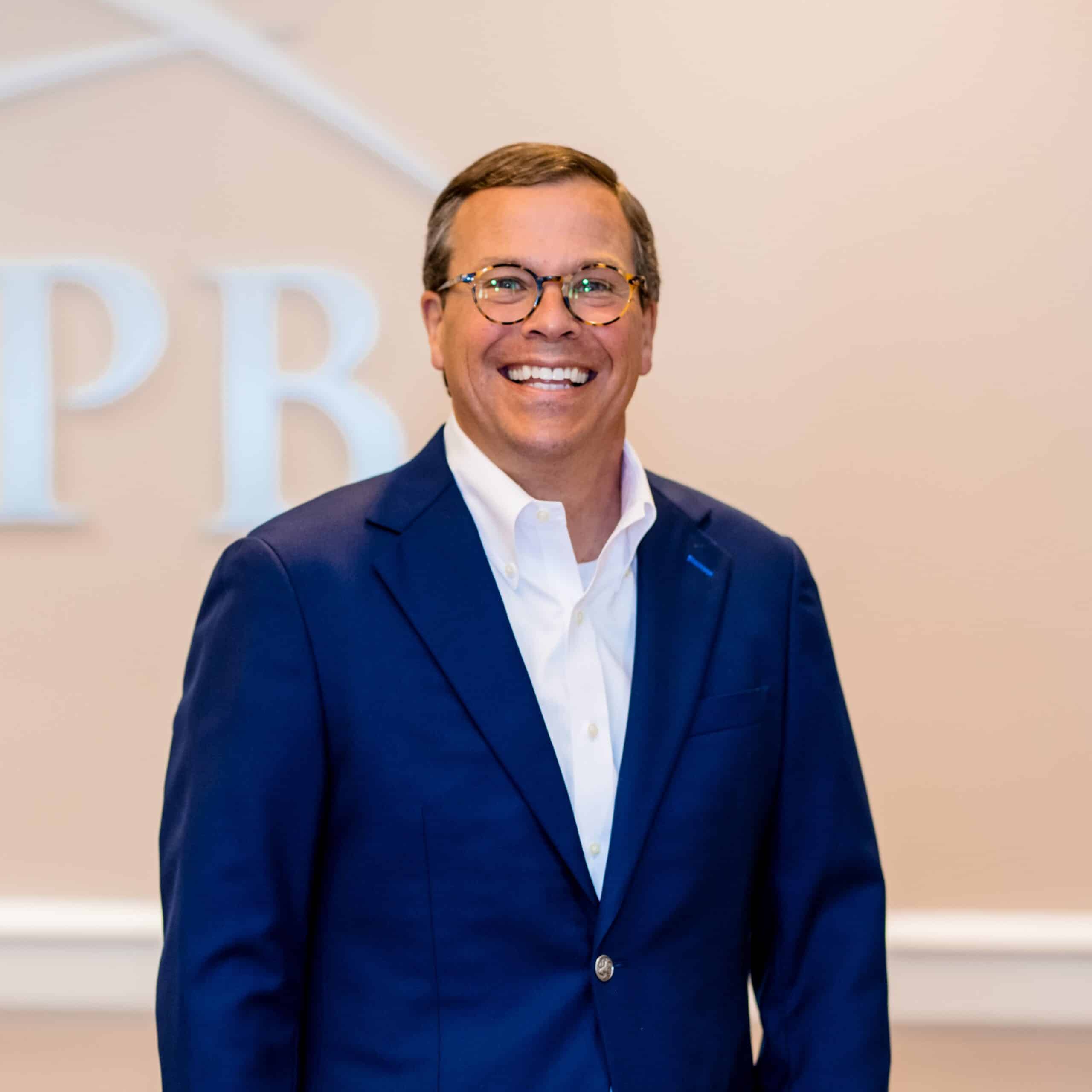 A man with glasses in a blue suit jacket over a white shirt smiles indoors, with blurred initials PB visible in the background.