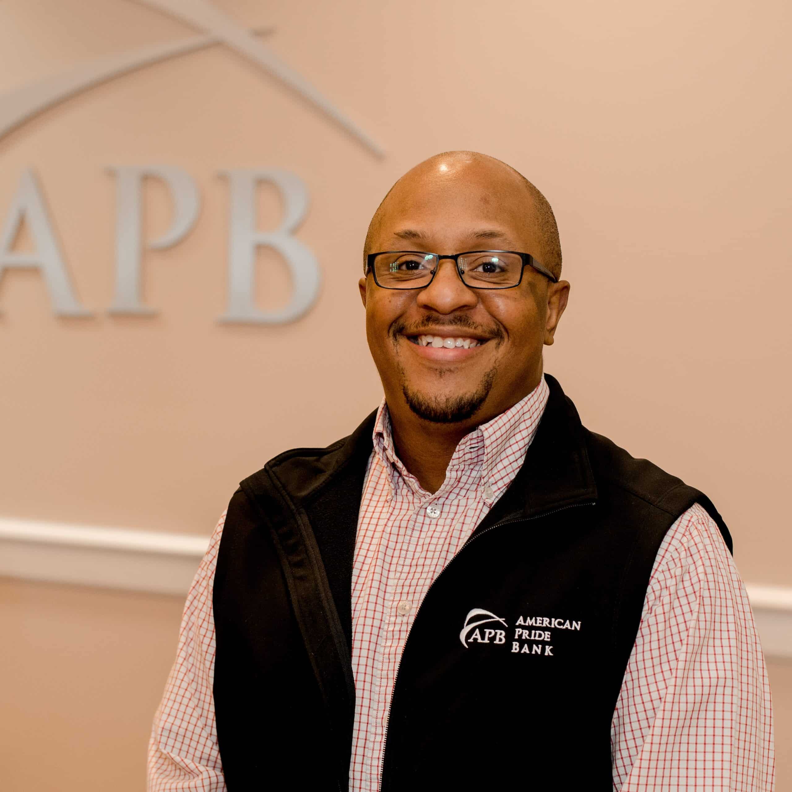 A man wearing glasses and a black American Pride Bank vest smiles at the camera, standing in front of a wall with the APB logo.
