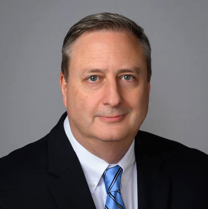 A middle-aged man with short, neat brown hair in a black suit jacket, white shirt, and blue striped tie poses against a gray background.