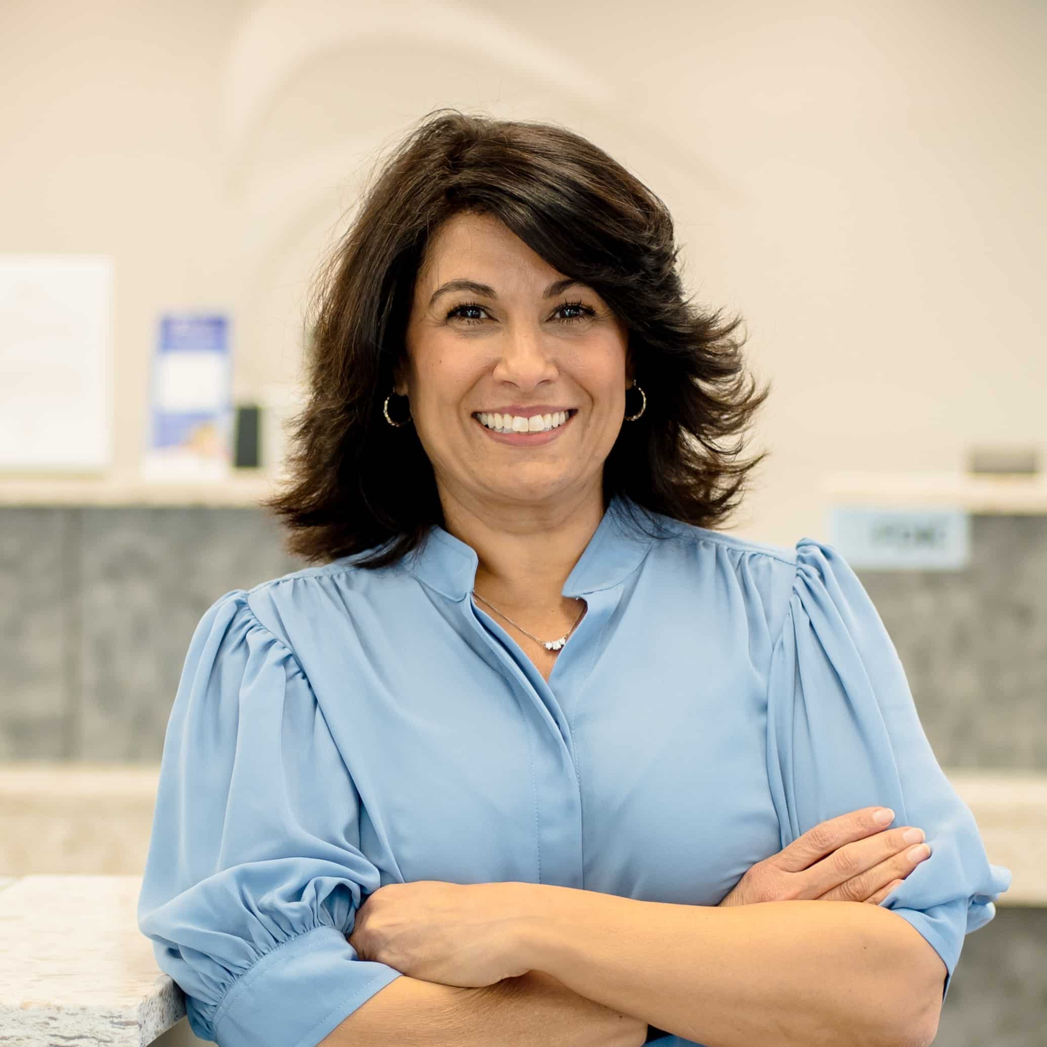 A woman with shoulder-length dark hair in a light blue blouse smiles confidently with arms crossed in a bright modern office.