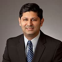 A man with dark hair in a dark suit, light blue shirt, and striped tie smiles at the camera in front of a plain gray background.