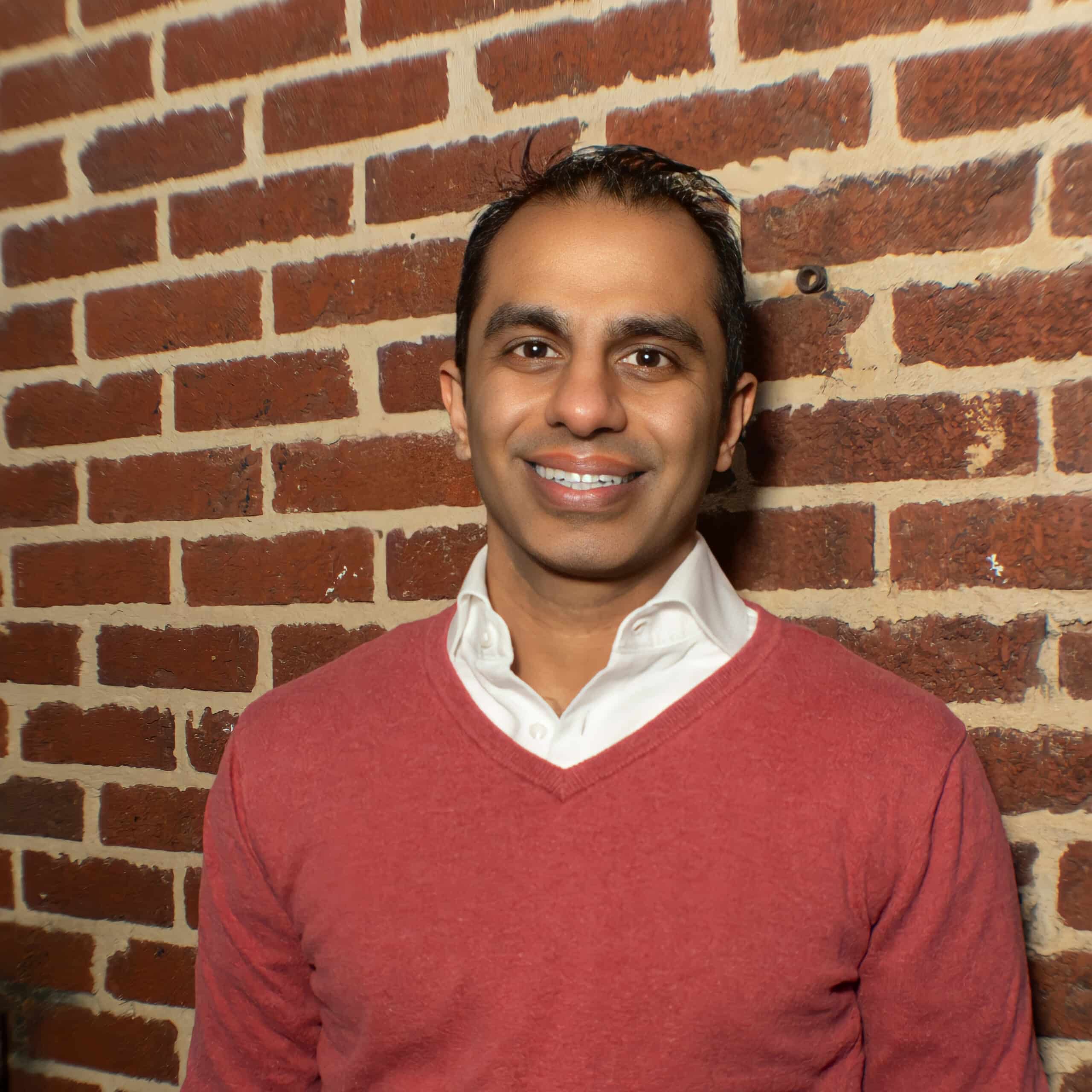 Man wearing a red sweater and white collared shirt smiles while standing in front of a brick wall.